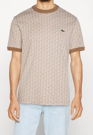 Homme portant une chemise beige à manches courtes à motifs avec une bordure brune et un logo crocodile vert sur la poitrine, associée à un jean bleu clair.