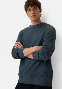 Granatowy sweter z okrągłym dekoltem, ściągaczami przy mankietach i u dołu, z małą metką z logo na piersi oraz fakturowanym materiałem.