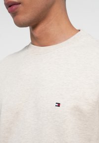 Maglione beige chiaro con colletto rotondo. Presenta un piccolo logo ricamato in blu navy, rosso e bianco sul lato sinistro del petto. Tessuto morbido.