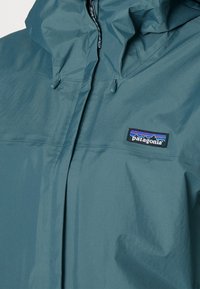 Teal vattentät jacka med huva, dragkedja och justerbara dragsnören. Innehåller en Patagonia-logotyp på bröstet.