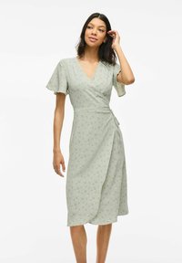 Robe verte avec des manches courtes et aérées, présentant un motif floral subtil et un détail à nouer à la taille. Tissu doux et texturé.