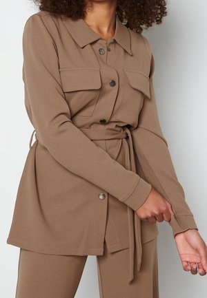 Femme portant une veste marron ceinturée avec des boutons en métal et deux poches à rabat sur la poitrine, ajustant sa manche.