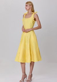 Robe midi jaune avec encolure carrée, fines bretelles nouées en nœuds, tissu texturé, jupe évasée et sandales blanches à lanières.