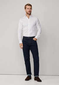 Biela pruhovaná košeľa s dlhými rukávmi, skombinovaná s tmavo modrými džínsami. Hnedé mokasíny a modré ponožky viditeľné na členkoch.