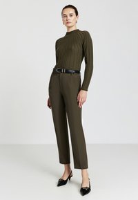 Top lungo a maniche lunghe verde oliva a costine con colletto alto, abbinato a pantaloni olive sartoriali. Cintura nera e décolleté neri a punta completano il look.