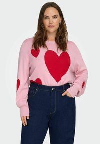 Růžový svetr s velkými červen ými srdíčkovými vzory, žebrovaným kulatým výstřihem a manžetami, kombinovaný s tmavými džínami s vysokým pasem.