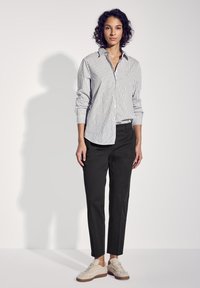 Gestreepte overhemdbloes in wit en marineblauw, gecombineerd met zwarte taps toelopende broek en beige gestructureerde sneakers. Strakke lijnen, casual pasvorm.