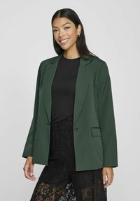 Grønn blazer med skreddersydd design, notklapp, enkel knappelukking og to frontlommer, brukt over en svart topp og blondebukser.
