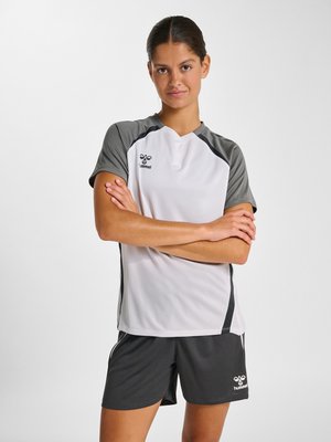 Ung kvinde står med armene krydset iført hvid og grå Hummel sportstrøje og mørkegrå shorts mod en ensfarvet baggrund.