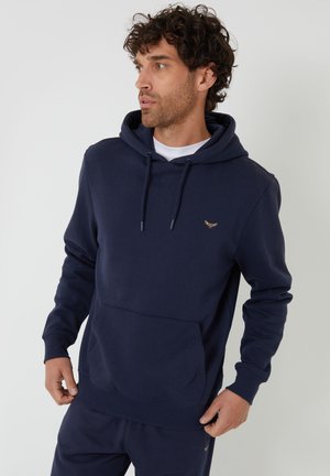 Uomo con capelli ricci che indossa una felpa con cappuccio blu navy e pantaloni coordinati, in piedi contro uno sfondo chiaro e semplice, che guarda di lato.