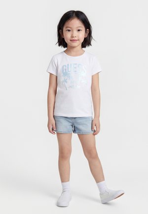 Young girl with short dark hair wearing white graphic t-shirt, light denim shorts, white socks, and white slip-on shoes standing on white background.