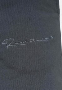 Grijze stof met een ribbeltextuur, voorzien van een geborduurd scriptlogo in lichter grijs, prominent gepositioneerd op het kledingstuk.