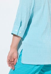 Blusa a righe blu chiaro con maniche arrotolate, dettagli con bottoni bianchi e spacchi laterali. Il materiale appare liscio con una consistenza fresca, abbinata a pantaloni coordinati.