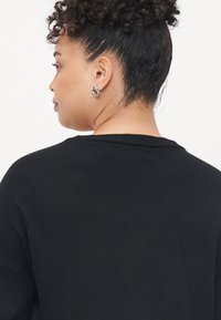 Pulover negru cu un model texturat de tricot, având o gâtă largă și o croială lejeră. Cercei rotunzi argintii vizibili pe urechea modelului.