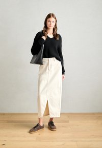Pull en tricot côtelé noir, jupe crème jusqu'au genou avec fente à l'avant, et baskets léopard imprimé brun. Sac fourre-tout noir tenu dans la main gauche.