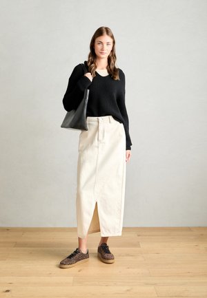 Schwarzer gerippter Strickpullover, cremefarbener knielanger Rock mit Frontschlitz und braune Sneaker im Leopardenmuster. Schwarze Tote-Bag in der linken Hand gehalten.