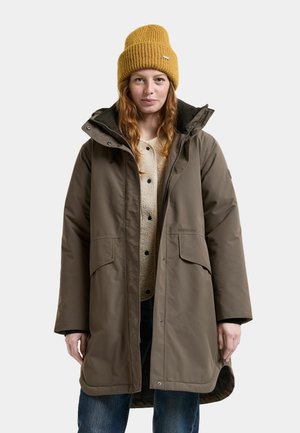 Jeune femme portant un bonnet en maille moutarde, un parka marron avec de grandes poches, une veste polaire beige et un jean bleu, debout devant un fond uni.