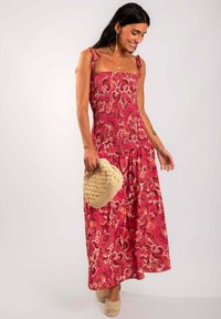 Vestido maxi rojo con estampado floral, bodice ajustado, textura fruncida y tirantes ajustables. Combinado con un bolso de mimbre y alpargatas ligeras.
