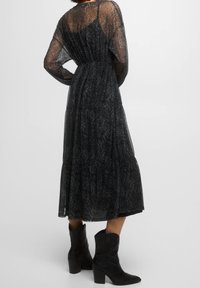 Robe noire transparente avec un motif paisley, manches longues, taille cintrée et jupe à volants ; portée avec des bottes à talons noires.