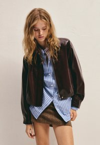 Tânără cu păr blond ondulat, îmbrăcată cu o jachetă din piele maro închis, cămașă în dungi albastre și fustă mini cu model maro, stând în fața unui perete simplu.