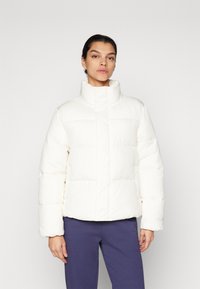 Pufferjack in off-white, met een hoge kraag, gestikte ontwerp en elastische manchetten. Het materiaal lijkt glad en gewatteerd voor warmte.