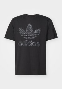 Svart bomullströja med en stor texturerad grå och svart Adidas-logotyp och tre bladdekorationer på bröstet. Rund halsringning och korta ärmar.