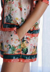 Zestaw loungewear z jedwabiu w kwiatowy wzór, składający się z bluzki z rękawami i szortów, w intensywnych różowych i zielonych kolorach, podkreślony koralowymi wykończeniami i kieszeniami.
