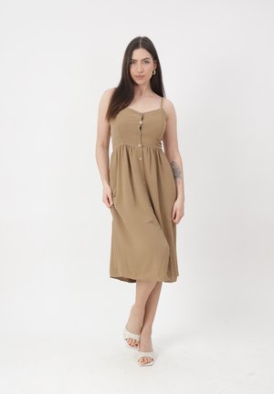 Ung kvinde med langt mørkt hår iført en beige midi-kjole med knapper og hvide åbne hæle, stående mod en ens hvid baggrund.