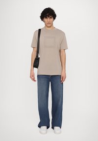 Beige bomulds t-shirt med et firkantet motiv på forsiden, parret med løstsiddende blå denimjeans og hvide sneakers.