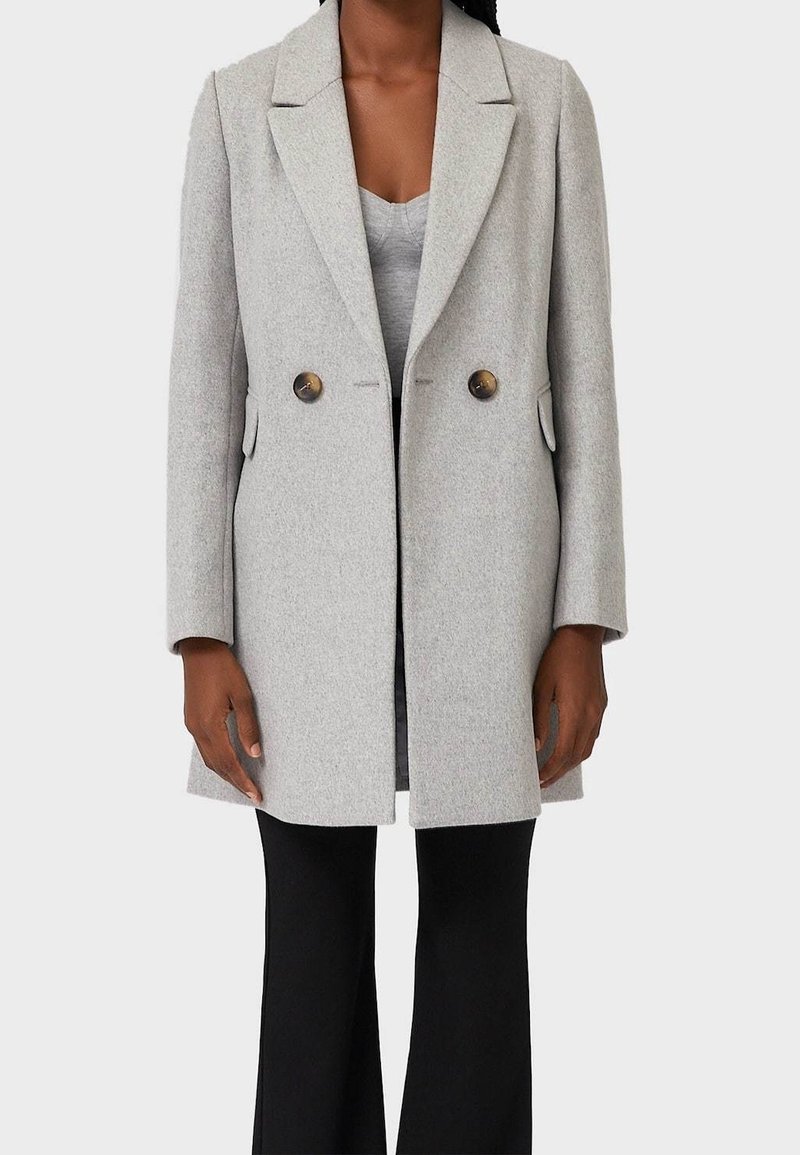 Femme portant un manteau en laine gris clair à double boutonnage sur un haut gris et un pantalon noir, debout devant un fond uni.