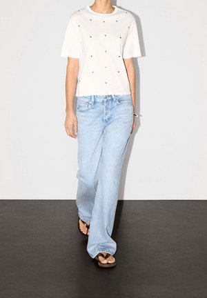 Osoba ubrana w biały krótki top z małymi ozdobami, jasnoniebieskie szerokie jeansy oraz czarne klapki, idąca do przodu.