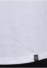 Camiseta de algodón blanca con una textura suave, mangas cortas, dobladillo curvado y una pequeña etiqueta negra con el logotipo en el lado inferior.