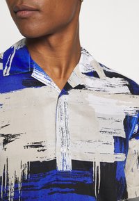 Skjorta med krage, abstrakt penselmönster i blått, svart och beigt; tillverkad av lättviktsmaterial; knäppning med knappar.