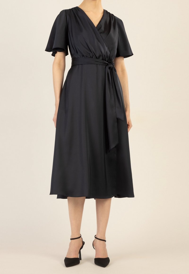 Femme portant une robe midi portefeuille noire avec des manches courtes volantées et une taille nouée, associée à des escarpins noirs à bride cheville, debout sur un sol beige.