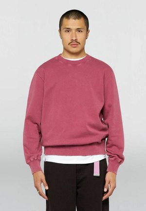 Uomo con i capelli corti e baffi che indossa una felpa rosa sbiadita sopra una camicia bianca e pantaloni neri con passante per cintura rosa.