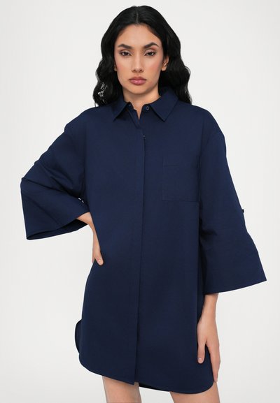 Abito camicia blu navy realizzato in un tessuto morbido e leggero; presenta una chiusura frontale con bottoni, un colletto a punta, maniche corte e larghe, e una tasca sul petto.
