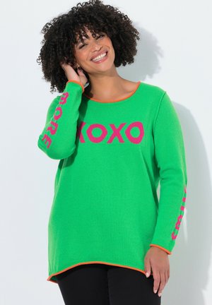 Smilende kvinde med krøllet hår iført en grøn sweater med lyserøde "XOXO" på brystet og "MORE" på ærmerne, sweateren har orange kant.