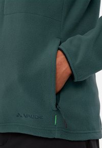 Pullover in pile verde con tasca con zip, superficie texturizzata e logo ricamato. Presenta una vestibilità casual e bordi neri a contrasto.