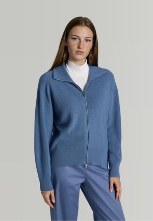 Vrouw met lang bruin haar die een blauwe vest met rits draagt over een wit topje en blauwe broek, staand tegen een effen lichtgrijze achtergrond.
