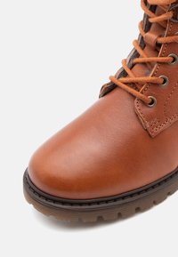 Bottines en cuir marron à lacets avec semelle en caoutchouc épaisse et œillets en métal, montrées en gros plan sur fond blanc.