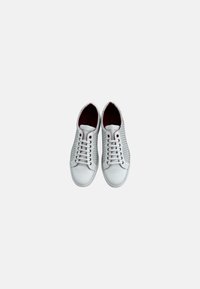 Zapatillas blancas con un panel texturizado, cordones planos, forro interior rojo y diseño de punta redondeada. Presentan una estética minimalista y un aspecto casual.