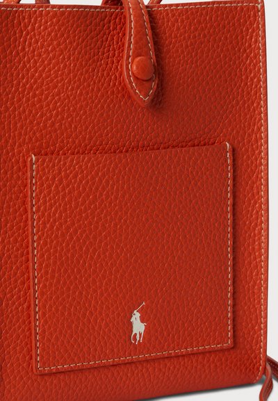 Červená kožená taška s texturovaným povrchem, přední kapsou a stříbrným logem. Taška má kontrastní bílé švy.