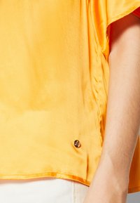 Top corto giallo in satin con una texture leggermente lucida, caratterizzato da un piccolo bottone nero sul bordo. Accentuato da linee pulite.