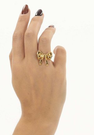 Hand met bruin en donkerpaars gelakte nagels, die een glanzende gouden strikvormige ring aan de middelvinger draagt, tegen een witte achtergrond.