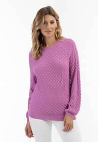 Texturiertes pinkes Sweatshirt mit muschelähnlichem Muster, runder Ausschnitt und langen Ärmeln. Das Material wirkt weich und wird mit weißen Hosen kombiniert.