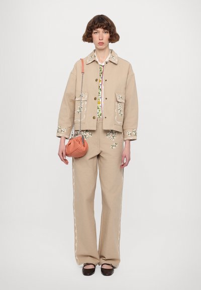 Giacca di cotone beige con ricami floreali, abbinata a pantaloni coordinati e una blusa bianca con una stampa floreale colorata. Piccola borsa arancione.