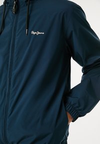 Chaqueta ligera de color azul marino con cierre de cremallera en el frente, puños elásticos y un pequeño logo en el pecho. El material parece suave y resistente al agua.