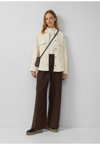 Crèmekleurige corduroy overhemdjasje met grote zakken, knoopsluiting aan de voorkant, gecombineerd met high-waisted bruine broek en chunky zwarte platformlaarzen.