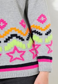Grå stickad tröja med livliga mönster i rosa, orange, grön och svart, inklusive stjärnor och zigzag-design, med ribbad mudd.
