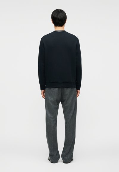 Άνδρας με την πλάτη στραμμένη, φορώνας μαύρο πουκάμισο μακρυμάνικο και χαλαρό γκρι παντελόνι, μπροστά σε μια απλή φωτεινή επιφάνεια.